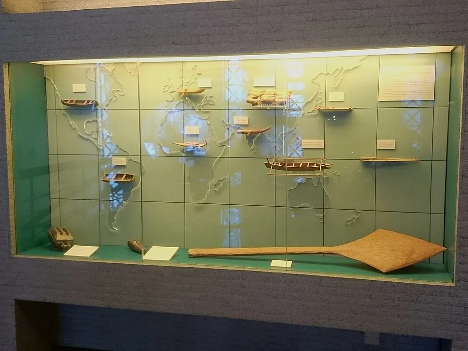 Models of differently shaped early boats and a wooden paddle in a museum case
